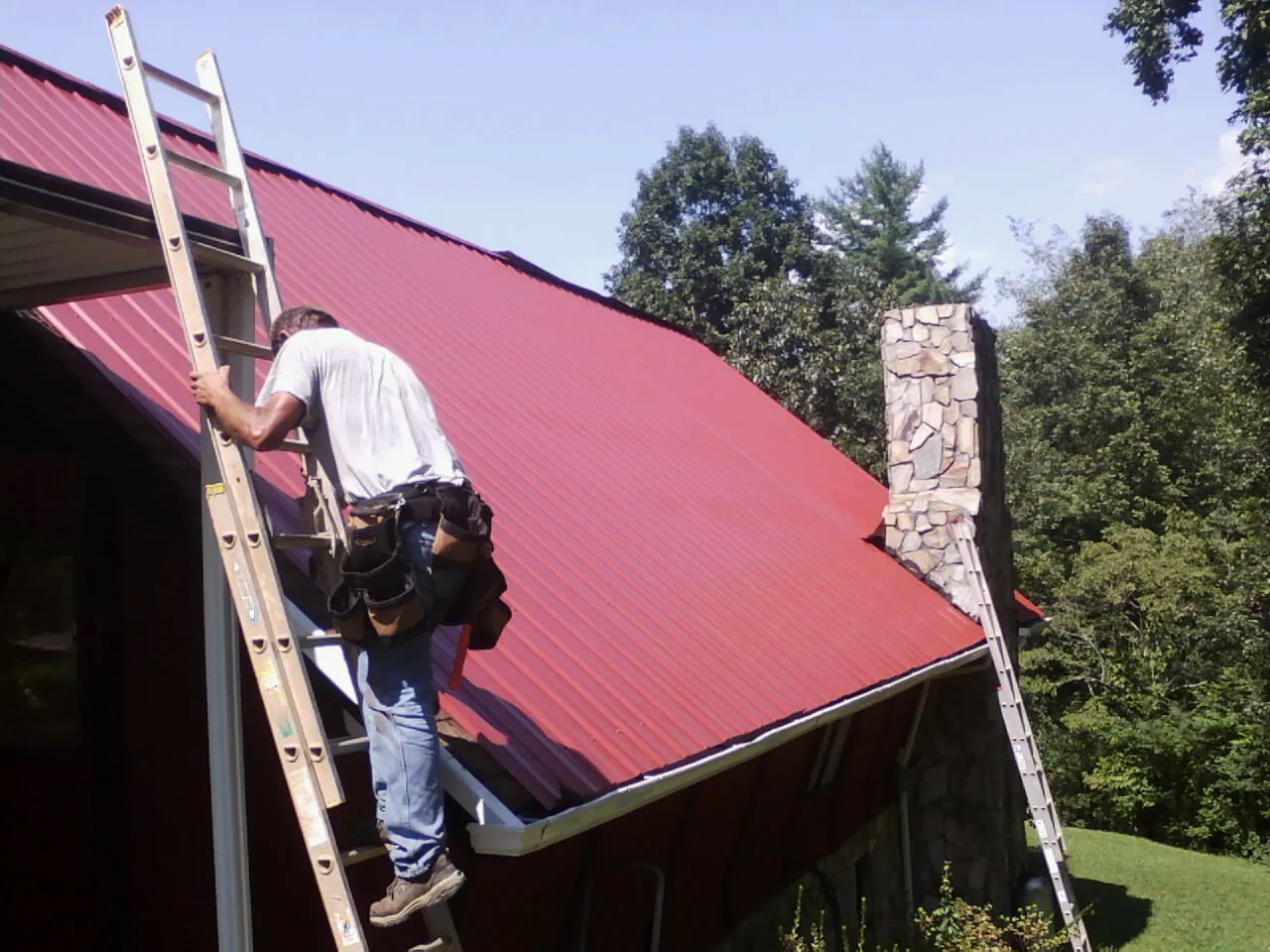 Roofer on a ladder beside a red metal roof and stone chimney — Soffit & Fascia Repair by Roofing Brothers in Mill City